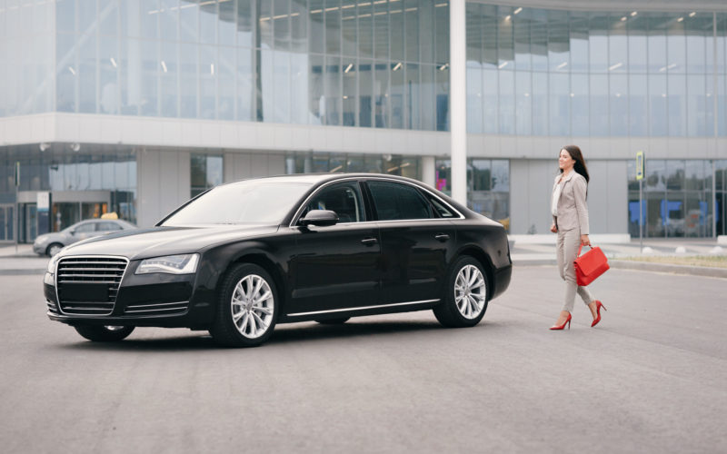 One Successful business woman walk toward a black car stopping on street with modern building as background. red leather bag in his hand.